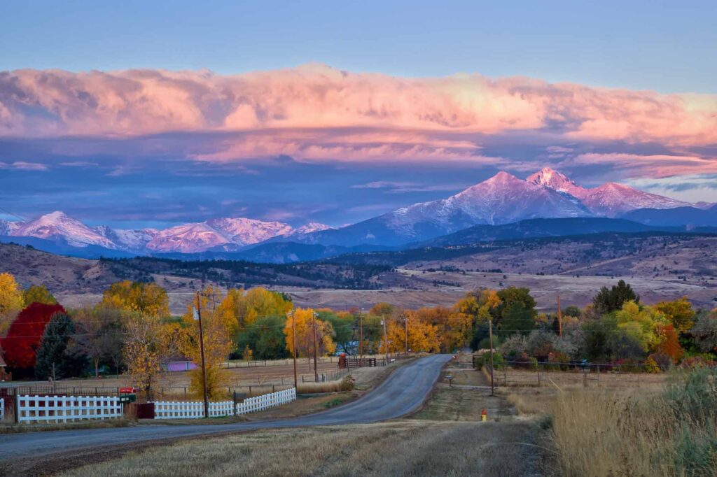 Long's Peak lights up at sunrise as a rural country road leads into the fall trees in Colorado